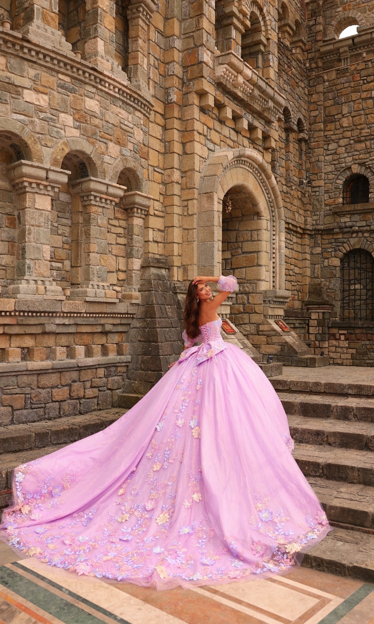 A woman in the Amarra 54234 Puff-Sleeve Quinceanera Dress, a long lavender gown with floral lace appliqués, poses on stone steps before a grand, ornate stone building.